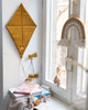 A cozy corner features a Velvet Kite Wall Decor in mustard, which is OekoTex®Standard100 certified, with bows hanging on the wall next to a white-framed window. Below, there is a stack of children's books, small knitted toys, and a soft blanket. A rainbow-shaped wall hanging and a stuffed animal decorate the children's room decor.