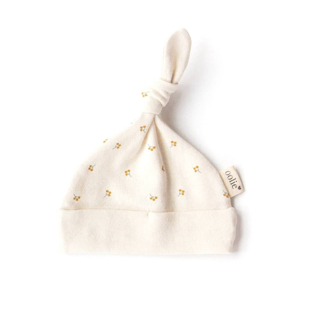 The Organic Baby Hat – Gold Sprig is a soft, cream-colored GOTS certified newborn hat made from organic cotton. It features a top knot, small yellow sprig prints, and a "oolie" fabric tag on the side, shown on a white background.