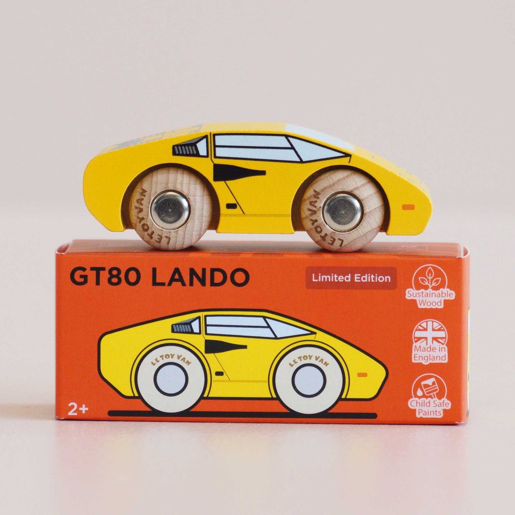 A bright yellow wooden No. 14 GT80 Lando supercar toy rests atop an orange box labeled “Made in England.” The packaging features icons for sustainable wood and a child-safe finish. Perfectly suited for ages 2 and up, this vibrant racing toy blends classic design with eco-friendly craftsmanship.