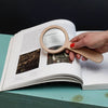 A hand with red nail polish holds the Wood Magnifier, made of beechwood, over an open book on a teal surface, enlarging text and images at 3x magnification.