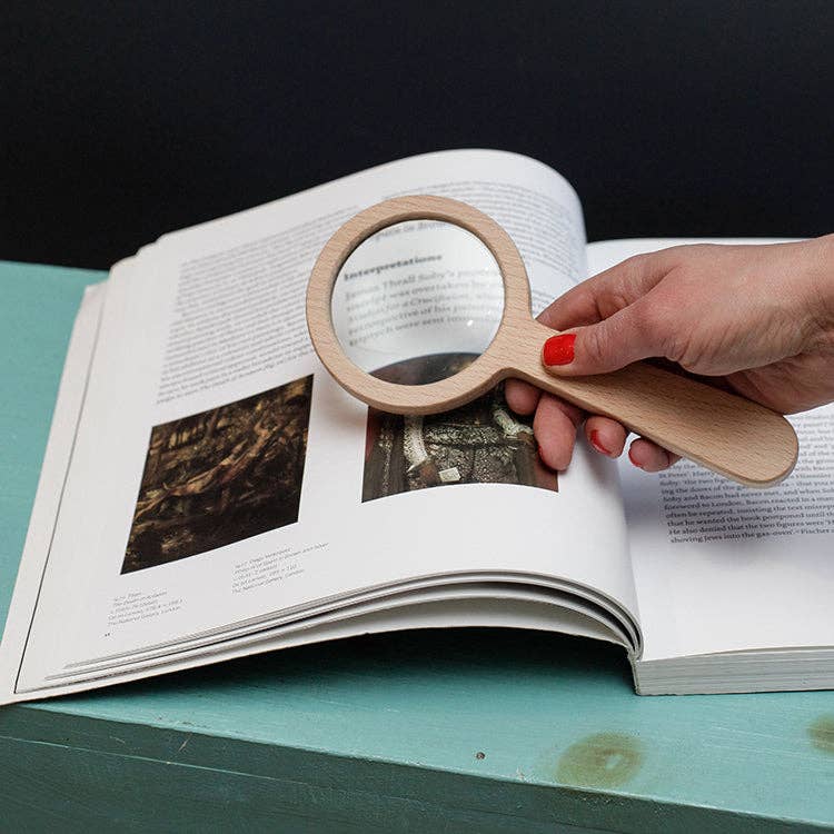 A hand with red nail polish holds the Wood Magnifier, made of beechwood, over an open book on a teal surface, enlarging text and images at 3x magnification.
