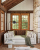 A cozy room featuring a white daybed adorned with Coral & Tusk Owl Family Tree Pillows and a green throw blanket. The daybed is positioned against a wooden wall with large windows, allowing natural light to stream in. A sleek floor lamp stands nearby, and a small side table holds books.