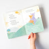 A child's hands hold open a Slumberkins Yak Kin + Lesson Book On Self Acceptance with illustrations of a happy fox and positive affirmations on a white background.