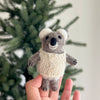 A hand holds a Baby Koala Finger Puppet/Ornament, featuring a small felt koala in a knitted cream sweater, displayed against a blurred green pine tree background.