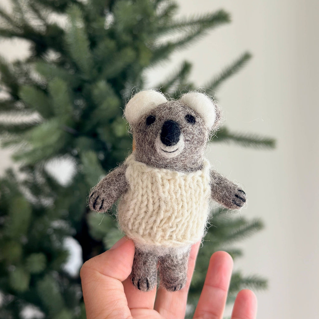 A hand holds a Baby Koala Finger Puppet/Ornament, featuring a small felt koala in a knitted cream sweater, displayed against a blurred green pine tree background.