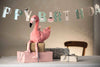The Steiff Flamingo Stuffed Animal sits atop wrapped gifts on a table with a "Happy Birthday" banner in the background, adding charm to the festive scene and a wooden chair partially visible.