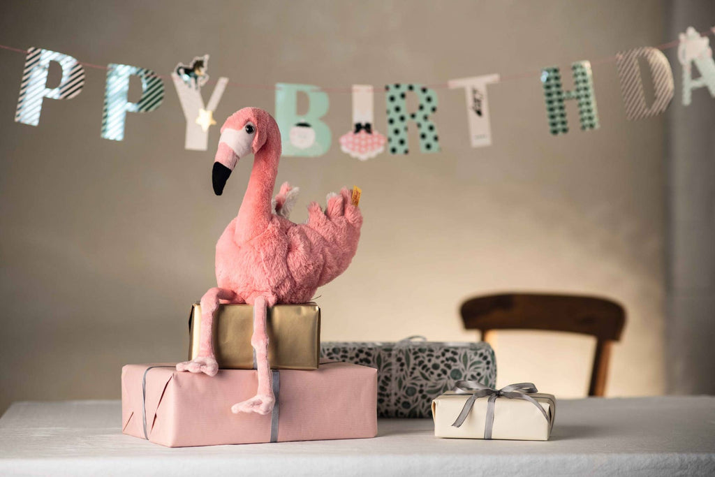 The Steiff Flamingo Stuffed Animal sits atop wrapped gifts on a table with a "Happy Birthday" banner in the background, adding charm to the festive scene and a wooden chair partially visible.