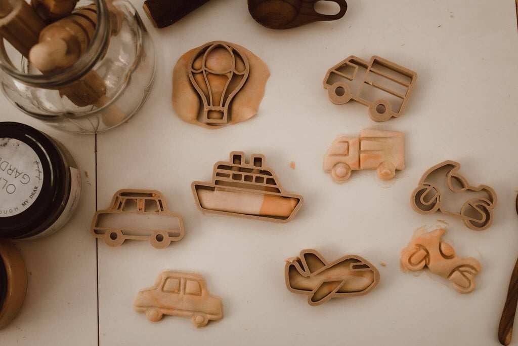 The Transport Cutter Set - Eco Playdough Tool features vehicle-shaped cutters (hot air balloon, ship, airplane, cars, bus, motorcycle, van) made from biodegradable PLA resting on a white surface alongside dough and jars—an ideal choice for creative play.