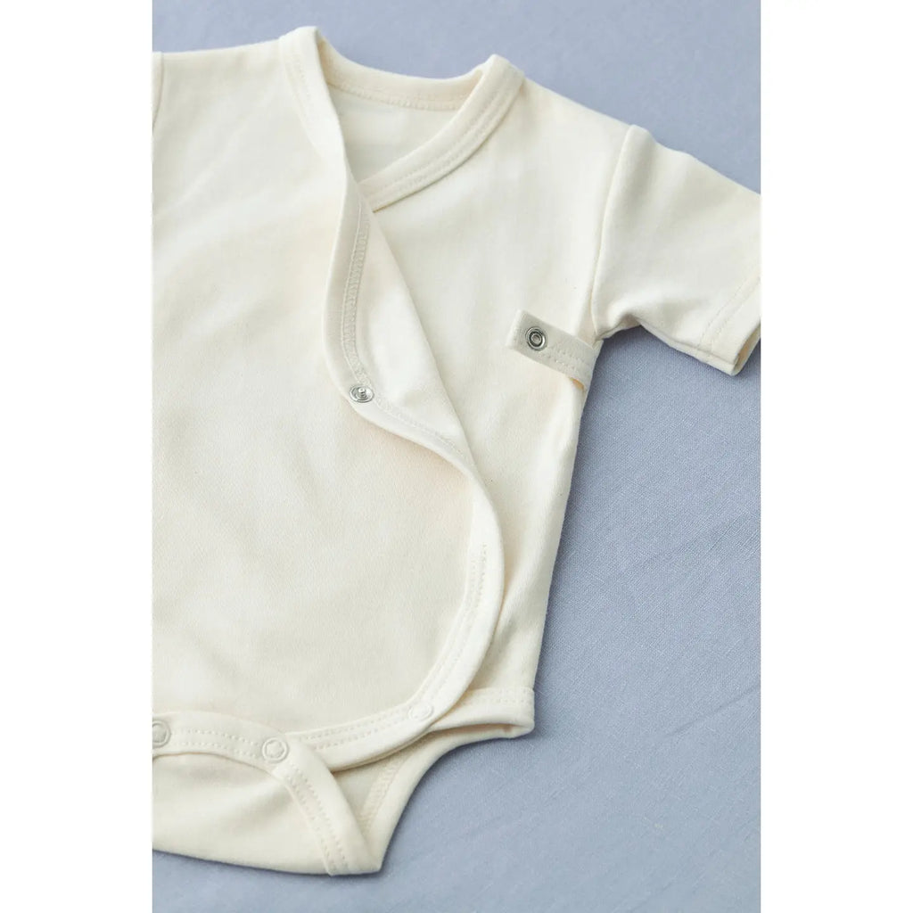 A cream-colored, organic cotton Baby Romper with short sleeves is displayed on a light blue surface. The romper features snap buttons for easy fastening, including two on the front and two between the legs, allowing for convenient diaper changes. It is also machine washable for effortless care.