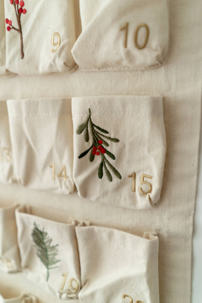 Close-up of the Sabo Concept Christmas Cotton Advent Calendar with gold-stitched numbers and botanical embroidery—red berries and green leaves—on pockets 14, 15, and 19. A charming holiday detail.