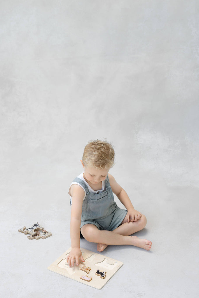 A young child sits on the floor wearing light blue overalls, playing with the Farm Animals Wooden Puzzle: A Tabitha Paige Puzzle. This adorable wooden peg puzzle, featuring charming farm animals, is designed for little hands and encourages learning through play. The background is plain and light-colored.
