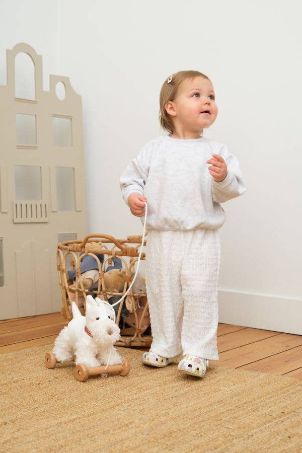 A toddler in white pajamas stands on a wooden floor, holding the string of Pull-Along Lola. Behind them are a wicker basket and a cardboard playhouse, inviting developmental play and supporting gross motor skills.
