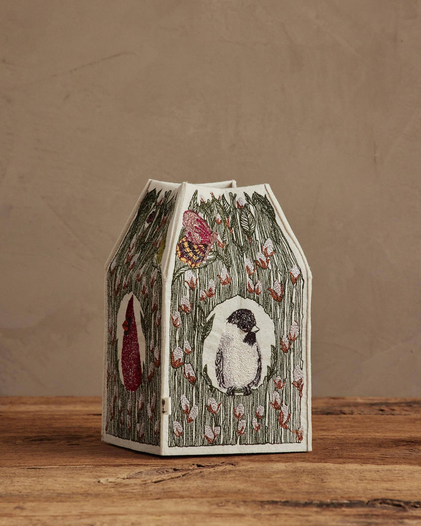 A paper lantern with embroidered bird and floral designs sits on a wooden surface beside the Coral & Tusk Birdhouse Tissue Box Cover, set against a plain beige background.