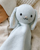 The Cuddle + Kind Organic Cotton Baby Bunny Lovey (Baby Blue)—a soft, blue bunny with floppy ears and a smiling face—rests beside a baby in cream knit clothing.