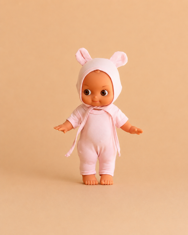 The Minikane Mini Pancha Doll - Ninon with Pale Pink Mouse Outfit stands on a beige background, facing forward with arms slightly outstretched. Crafted from high-quality vinyl, this charming figure inspires imaginative play for both children and collectors.