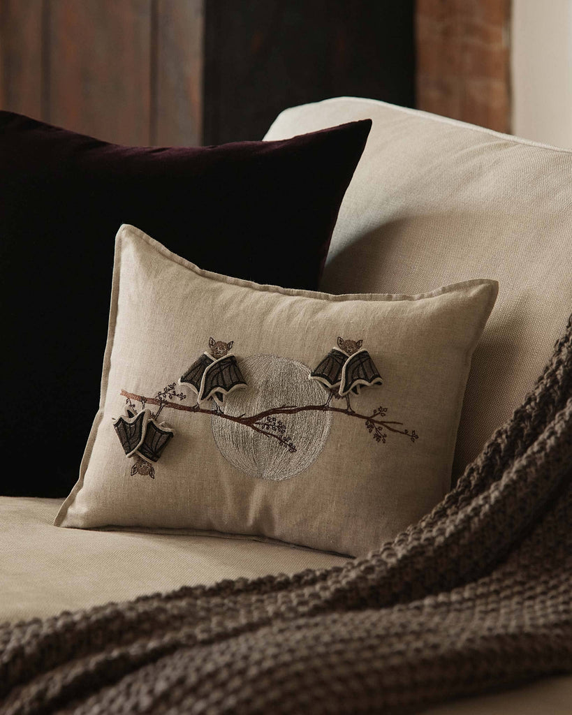 The Coral & Tusk Bat Branch Pillow, featuring embroidered black bats on beige linen, rests on a light couch beside a brown blanket and velvet cushion—an ideal subtle touch for Halloween décor. Ships in one week.