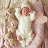 A sleeping baby in a fuzzy white outfit and hat lies on a pink blanket, cuddling the Cuddle + Kind Baby Honey Bear (Lavender Bow), with two hand-knit baby animals and a heart-shaped "My First Valentine's Day" sign nearby.