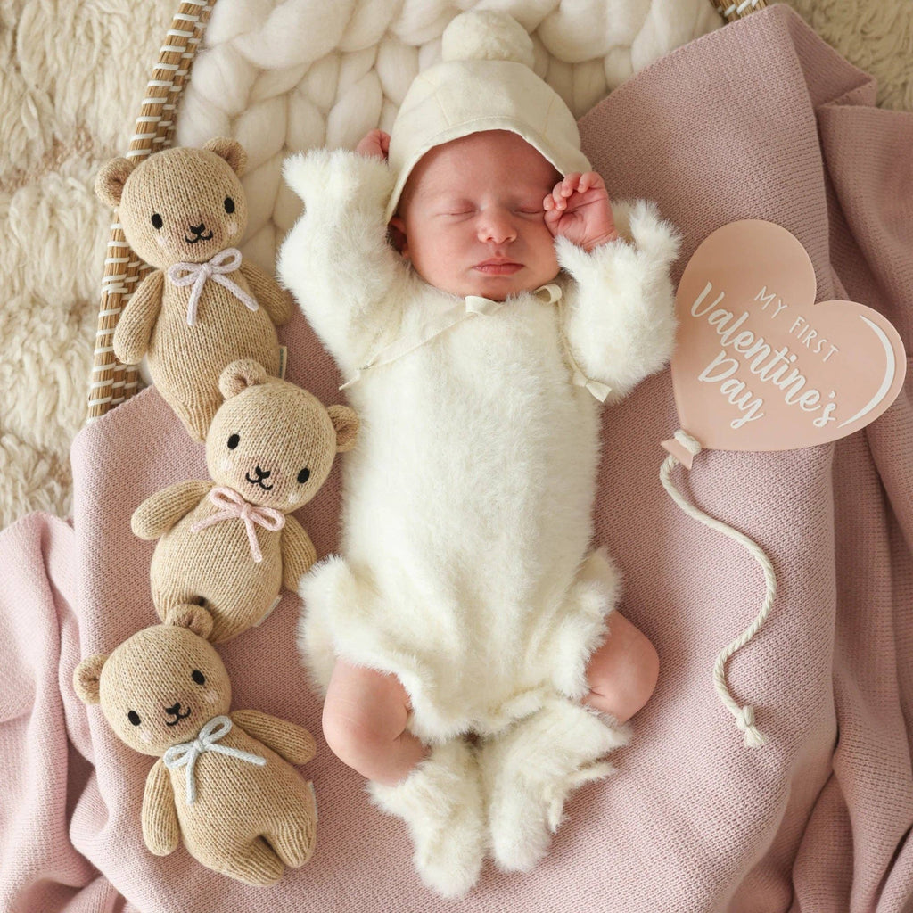 A sleeping baby in a fuzzy white outfit and hat lies on a pink blanket, cuddling the Cuddle + Kind Baby Honey Bear (Lavender Bow), with two hand-knit baby animals and a heart-shaped "My First Valentine's Day" sign nearby.