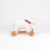 Pull-Along Lola is a white plush dog toy with a red checkered collar, set on a wooden base with four wheels and a pull string to encourage gross motor skills and developmental play.