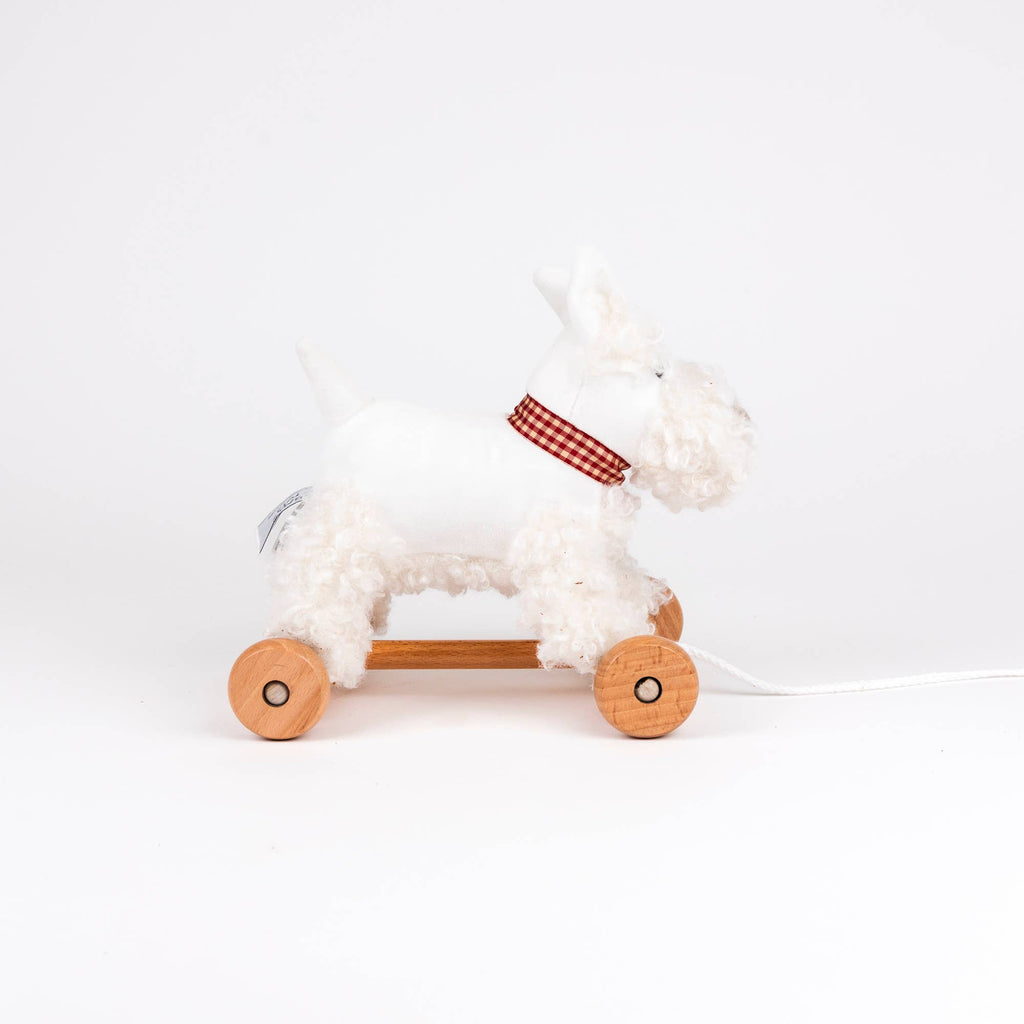 Pull-Along Lola is a white plush dog toy with a red checkered collar, set on a wooden base with four wheels and a pull string to encourage gross motor skills and developmental play.