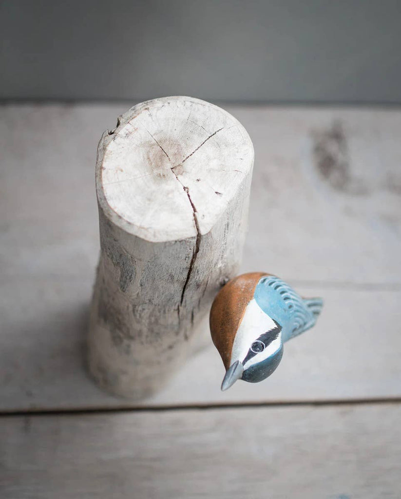 The Hand Carved Ornament - Red Breasted Nuthatch Bird (8"H) features a colorful, handmade wooden figurine perched sideways on a light, vertical sustainable softwood log, displayed on a pale wooden surface with a simple background.