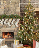 The living room is inviting with its stone fireplace, decorated with six embroidered stockings. Beside a stack of wrapped gifts stands a beautifully decorated Christmas tree, adorned with lights and ornaments, featuring a small Rudolph Ornament. The scene radiates warmth and holiday cheer, making it ideal for celebrating Christmas Day.