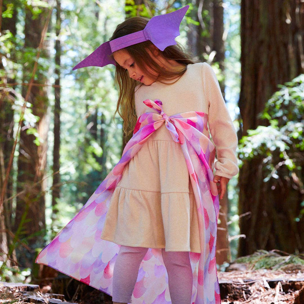 In a forest, a young girl wears a beige dress, lavender tights, a purple headpiece, and a pink and purple Sarah's Silks Magical Creatures Playsilk - Pink Unicorn cape tied at her waist as she looks down with trees behind her.