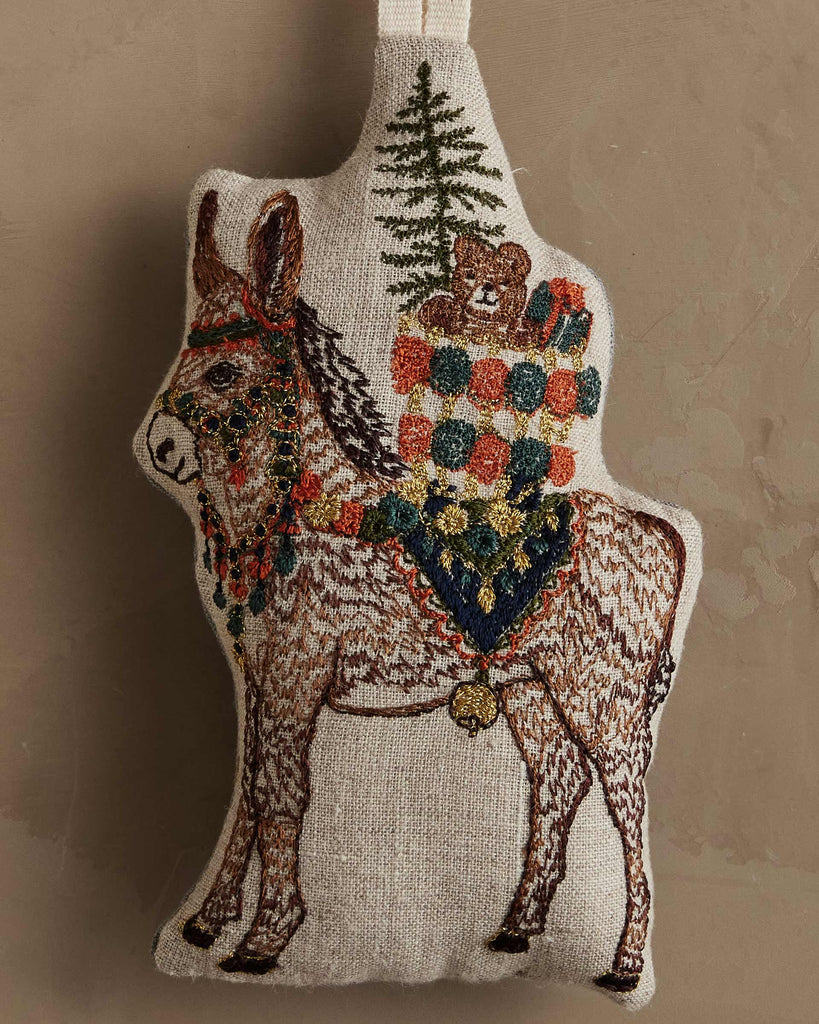 Coral & Tusk Christmas Donkey Ornament featuring an embroidered natural linen donkey carrying a stack of wrapped gifts and a small pine tree on its back.