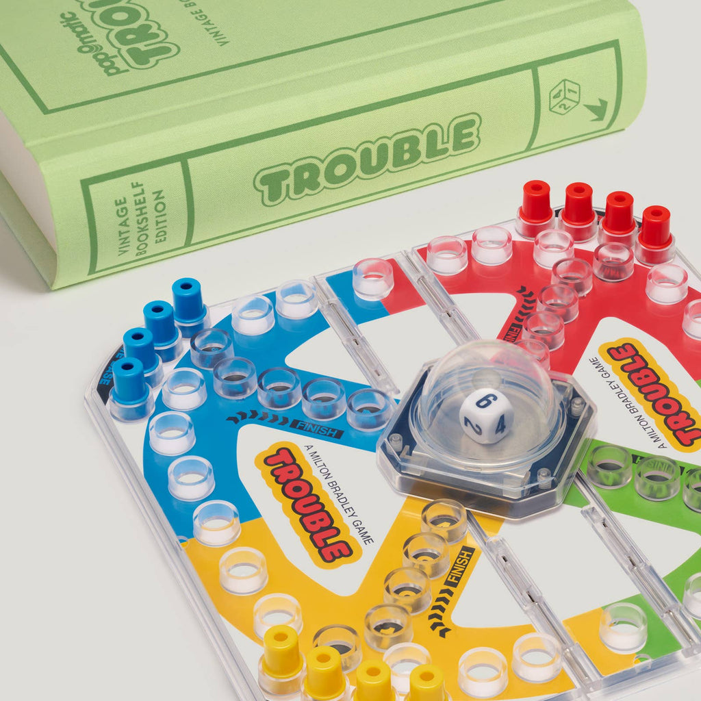 A close-up of the Trouble Vintage Bookshelf Edition game board shows colorful pegs and the Pop-O-Matic die roller, with the green book-shaped box labeled "Board Game: Trouble Vintage Bookshelf Edition" in the background.