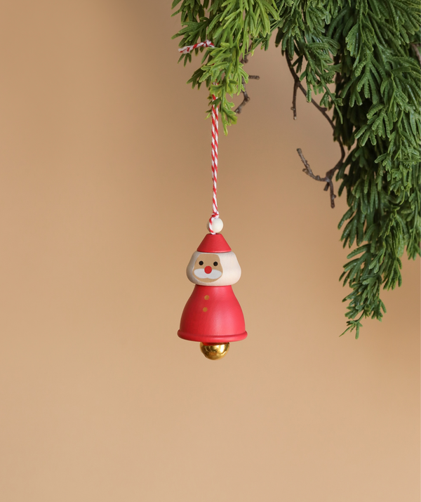 The Wooden Santa Ornament, featuring a red outfit and a white beard, hangs gracefully from a string on a green branch. Designed for decoration, this festive piece stands out beautifully against the plain, warm beige background.