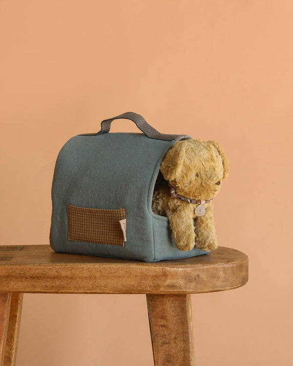 The Maileg Puppy in Dusty Blue Pet Carrier - Soft Sand rests partially inside its blue fabric carrier with a handle, placed on a wooden bench against a plain beige background.