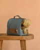 The Maileg Puppy in Dusty Blue Pet Carrier - Soft Sand rests partially inside its blue fabric carrier with a handle, placed on a wooden bench against a plain beige background.