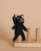 A small Black Cat Finger Puppet, crafted from needle-felted black felt with yellow eyes and pink ears, stands on a wooden toy featuring four vertical pegs. The background is a plain beige color.