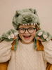 A smiling child in glasses and a chunky knit sweater poses playfully in the Wild & Soft Costume - T-Rex Dino: a plush green dinosaur outfit with paw-shaped mittens and a dinosaur headpiece featuring teeth.