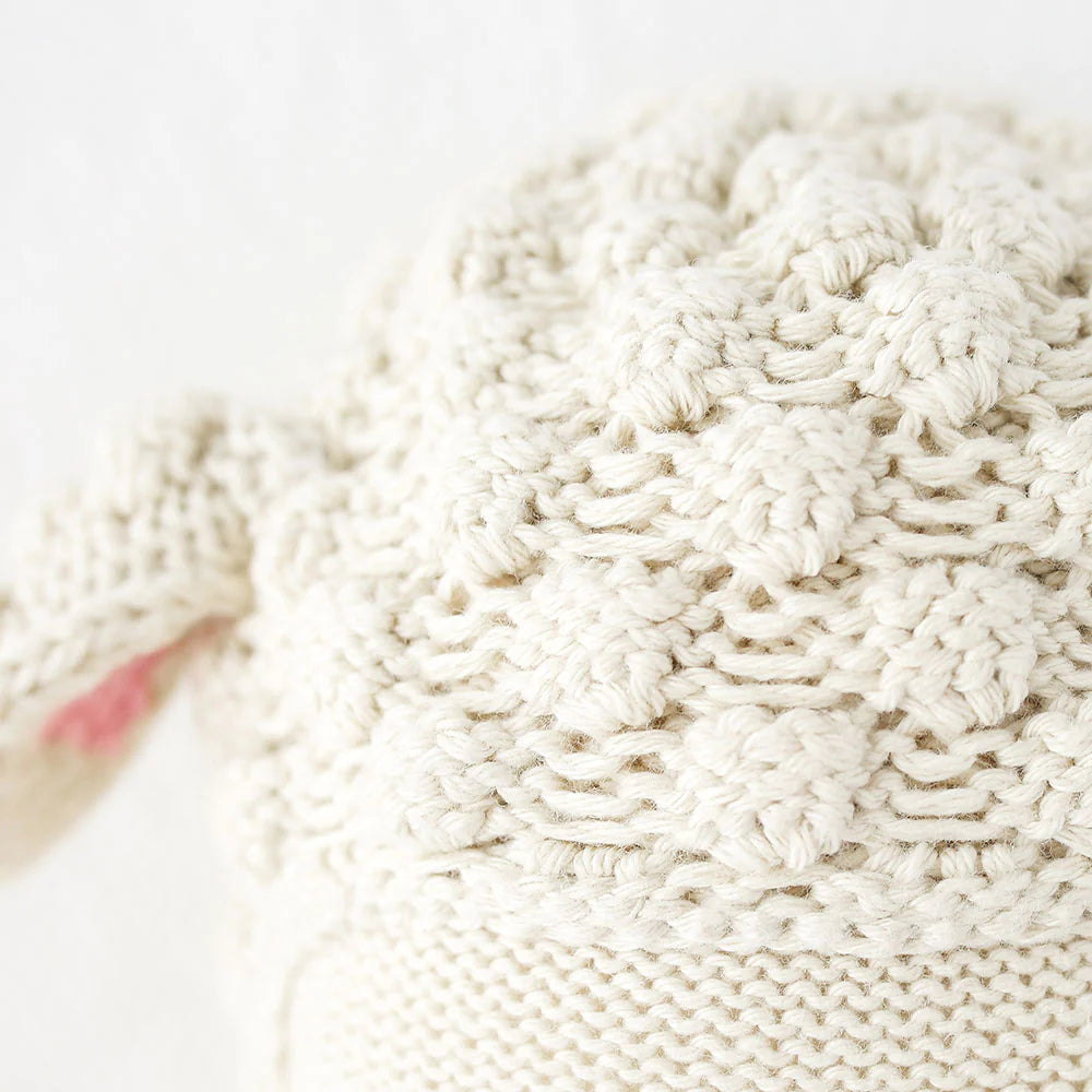 Close-up of a cream-colored, hand-knit Cuddle + Kind Lucy the Lamb doll with a raised bobble pattern and a touch of pink on the left side.