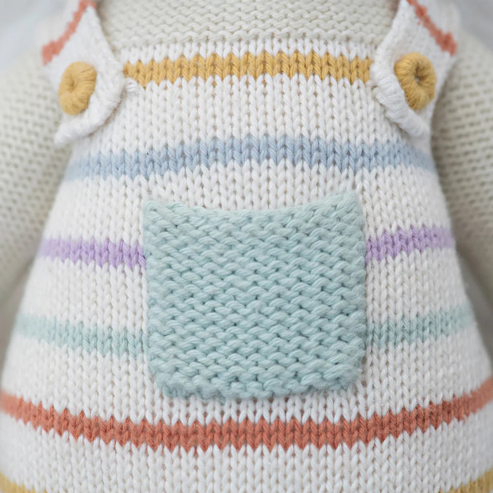 Close-up of Cuddle + Kind Avery the Lamb, a hand-knit doll in overalls with multicolored horizontal stripes, a light blue pocket, and yellow buttons—crafted as a sustainable, fair trade keepsake.