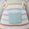 Close-up of Cuddle + Kind Avery the Lamb, a hand-knit doll in overalls with multicolored horizontal stripes, a light blue pocket, and yellow buttons—crafted as a sustainable, fair trade keepsake.