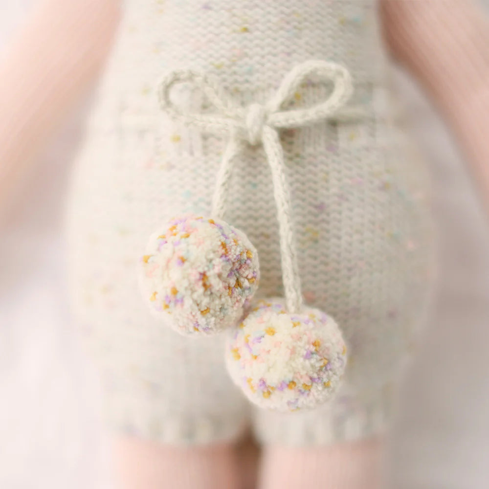Close-up of Cuddle + Kind Ella the Unicorn, a handmade doll in a light outfit with two fluffy pastel-speckled pom-poms at the waist. The background is softly blurred.