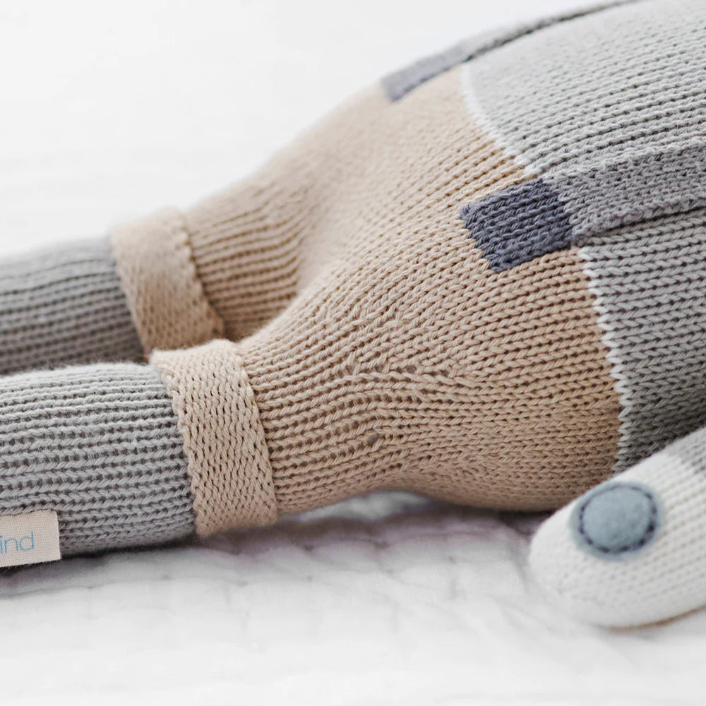 The Cuddle + Kind Noah the Dog, a fair trade hand-knit doll, rests on a white surface, displaying thoughtful craftsmanship and ethical sourcing.