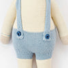 Cuddle + Kind Sebastian the Lamb is a sustainable fair trade hand-knit doll in light blue overalls with suspenders, cream-colored arms, legs, and torso. The doll’s head is not visible in the image.