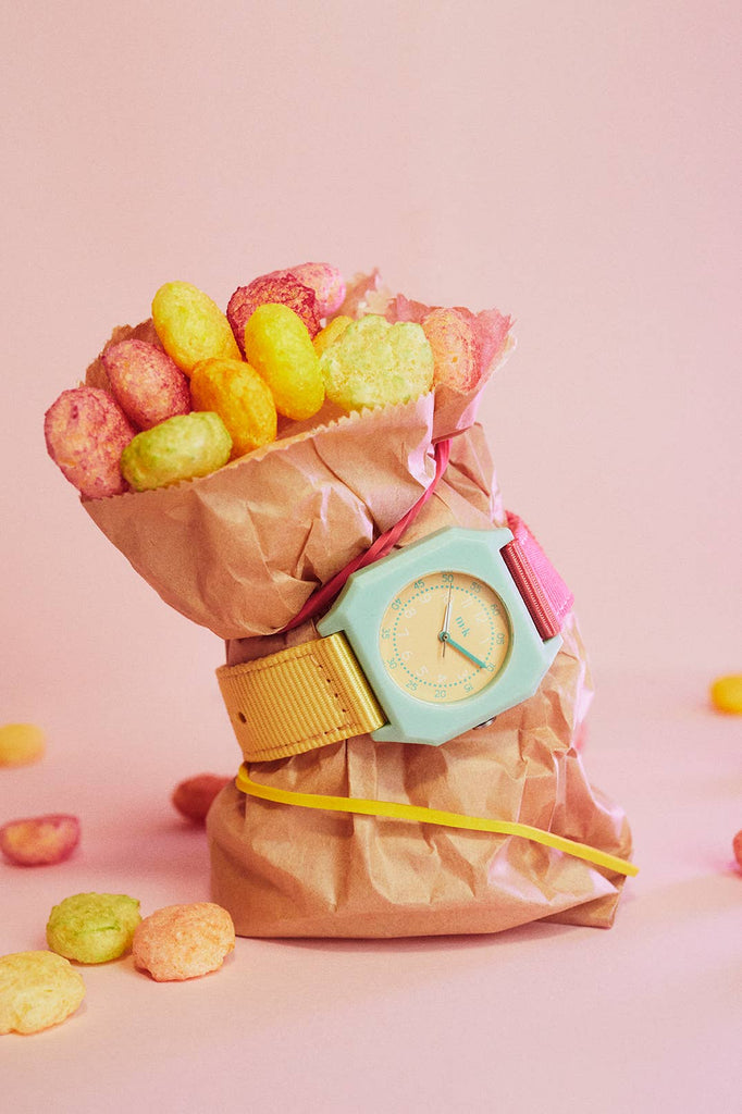 A brown paper bag filled with colorful round cereal pieces is wrapped with a Mini Kyomo Watch - Bubble Gum and secured with a pink rubber band, set against a pink background.