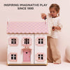 A young girl plays with Sophie's Wooden Dolls House, a pastel pink toy almost as tall as her. Above, the text reads: “Inspiring Imaginative Play Since 1995.” The softly lit, minimal setting highlights the charming design.