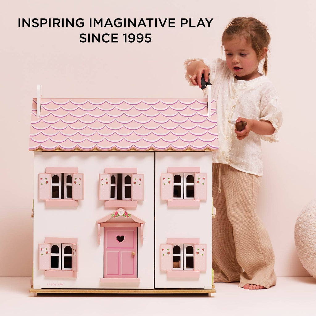 A young girl plays with Sophie's Wooden Dolls House, a pastel pink toy almost as tall as her. Above, the text reads: “Inspiring Imaginative Play Since 1995.” The softly lit, minimal setting highlights the charming design.