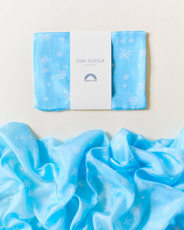 A blue silk fabric adorned with white snowflake patterns, partially folded and partly spread open, demonstrates its imaginative possibilities for creative play. The folded section bears a label stating "Sarah's Silk Giant Snow Playsilk" and features a small logo with rainbow lines.