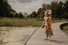 A child in the Costume - Tiger stands on a wooden boardwalk near a pond, surrounded by green plants and trees beneath a cloudy sky.