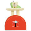 The Kitchen Weighing Scales is a wooden pretend play toy with a red base, cream top, and sliding dial from 0 to 4. Includes a green and white apple cut in half—perfect for kitchen role play fun.