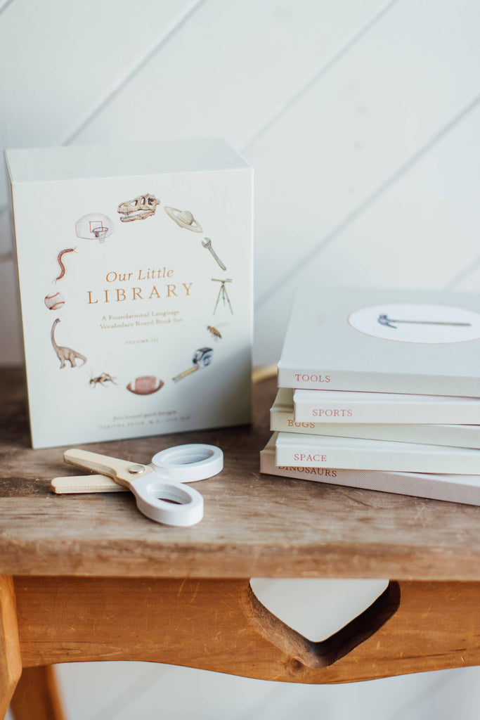 A box labeled "Our Little Library Vol. 3 (5 Board Books)" sits on a wooden bench beside titles "TOOLS," "SPORTS," "SPACE," and "DINOSAURS"—ideal for early language learning. A pair of white scissors rests nearby.