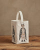 The Coral & Tusk Halloween Trick Or Treat Bag, cream-colored and small, features a bat with a pumpkin and a rabbit in a skeleton costume on the front. Shown on a wooden surface with a beige background. Ships in one week.
