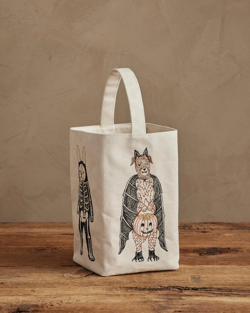 The Coral & Tusk Halloween Trick Or Treat Bag, cream-colored and small, features a bat with a pumpkin and a rabbit in a skeleton costume on the front. Shown on a wooden surface with a beige background. Ships in one week.