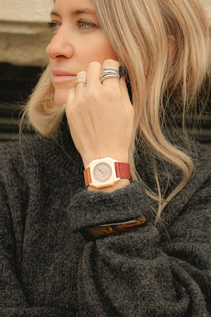 A woman with blonde hair wears a black sweater, several rings, and the Mini Kyomo Watch - Cherry Bomb with a brown leather strap, looking to the side and resting her hand near her face.